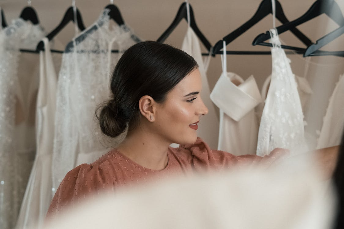 The image shows a woman with a soft bun hairstyle, admiring wedding dresses hanging in a boutique.