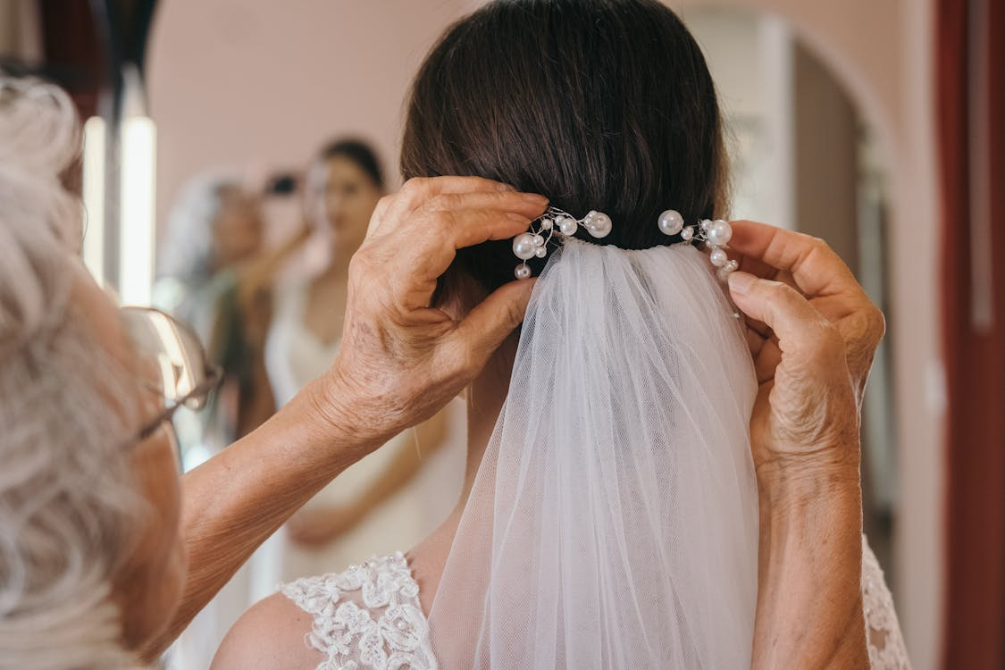 The image shows an older woman adjusting a bride's veil, with delicate pearl embellishments, in a dressing room.
