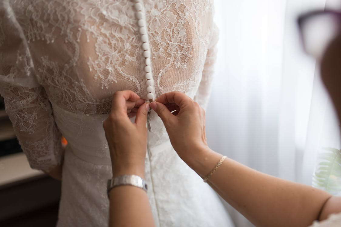 The image shows a close-up of a person helping a bride button up the back of her lace wedding dress.