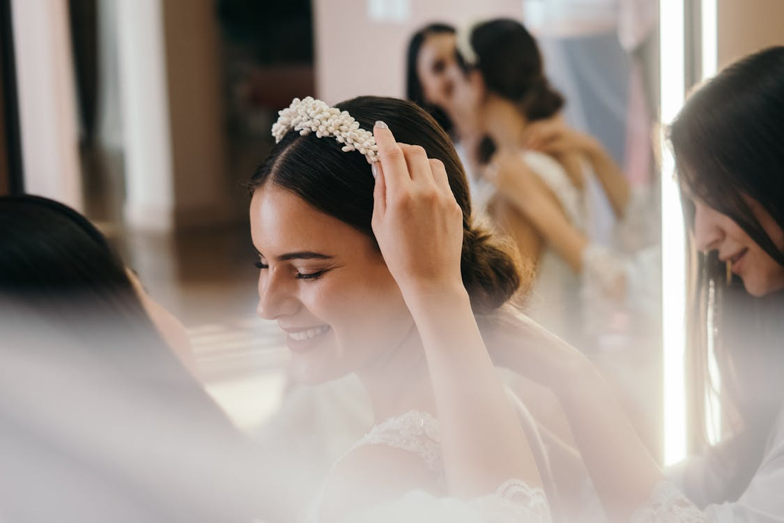 The image shows a bride smiling as a woman adjusts a pearl headband on her hair, with other bridesmaids in the background.